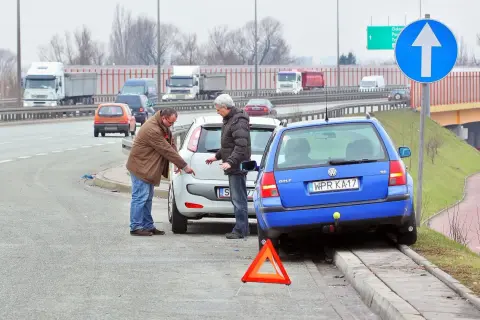 Dwóch mężczyzn dyskutuje przy uszkodzonych samochodach po kolizji. Trójkąt ostrzegawczy sugeruje, że może być spowodowanie kolizji mandat.