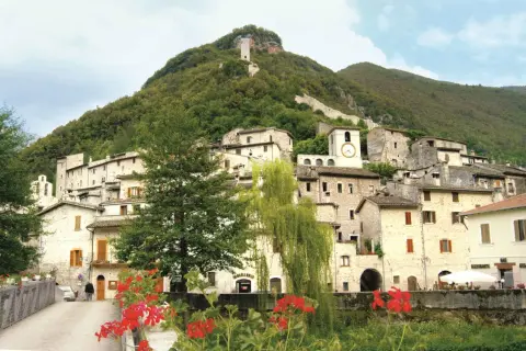 Antico borgo arroccato su un colle verdeggiante, uno dei paesi della Valnerina, con case in pietra, un campanile con orologio e una torre medievale in cima.