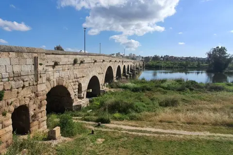 Puente romano de piedra con arcos sobre un río, bordeado de vegetación. Un camino de tierra serpentea por la orilla, evocando la antigua **Vía de la Plata**.