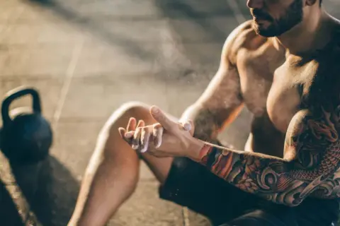 Ein Mann mit beeindruckenden herren tattoos bereitet sich mit Magnesium vor. Kettlebells liegen im Hintergrund.