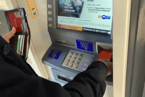 Osoba wkłada kartę do bankomatu, by wypłacić pieniądze. Widoczny portfel z kartami.