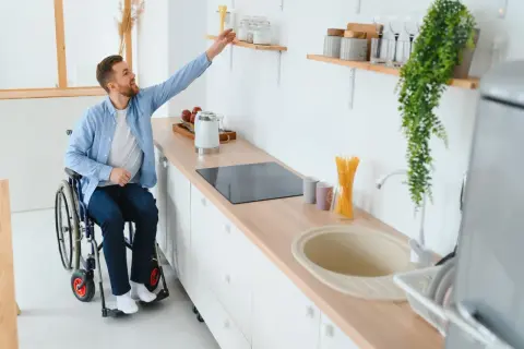 Un homme en fauteuil roulant attrape un objet sur une étagère dans une cuisine adaptée. Un exemple de priorité handicap logement social.