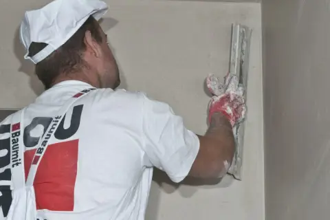Murarz w białej czapce i koszulce z logo Baumit nakłada tynk na ścianę za pomocą kielni.