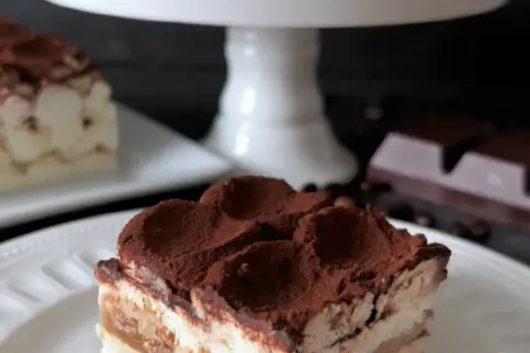 En bit krämig tiramisu, perfekt för ett tiramisu recept original. Pudrad med kakao, med kaffebönor och choklad i bakgrunden.