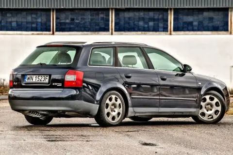Ciemnogranatowe Audi A4 B6 1.9 TDI Avant stoi na mokrym asfalcie przed budynkiem z niebieskimi oknami.