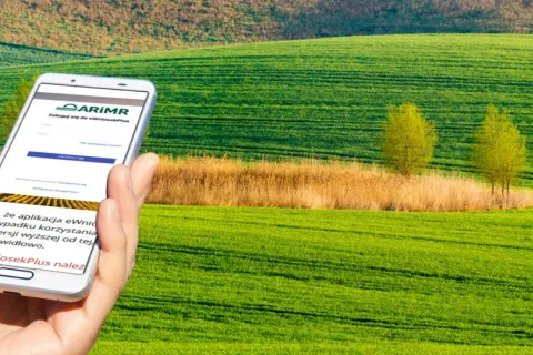 Dłoń trzyma smartfon z aplikacją ARiMR do składania wniosków o płatności bezpośrednie na tle zielonych pól.