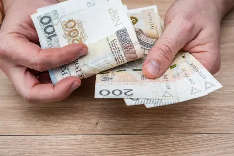 Dłonie trzymają polskie banknoty 100 i 200 zł. Może to być pomoc w opłaceniu rachunków, np. bon ciepłowniczy.