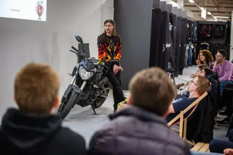 Mężczyzna w bluzie z płomieniami siedzi na motocyklu przed publicznością.
