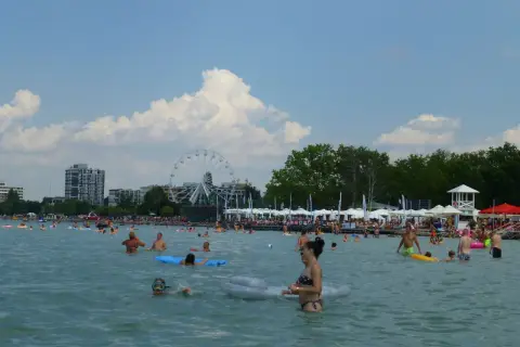 Ludzie kąpią się i relaksują na **Balaton plaża**. W tle widać diabelski młyn i budynki.