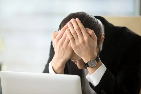 Pracownik w garniturze z rękami na twarzy, przy laptopie. Wygląda na przygnębionego, być może z powodu odpowiedzialności finansowej pracownika za błędy.