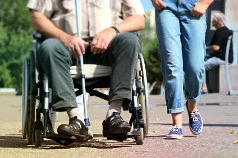 Persona in sedia a rotelle, con un evidente piede cadente invalidità, accompagnata da una persona che cammina al suo fianco.