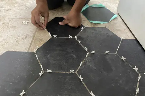 Hands place dark hexagonal tiles over existing floor tiles, using white spacers to ensure even gaps.
