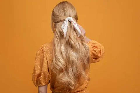 Chevelure blonde ondulée, coiffée en demi-queue avec un foulard fleuri, sur fond orange.