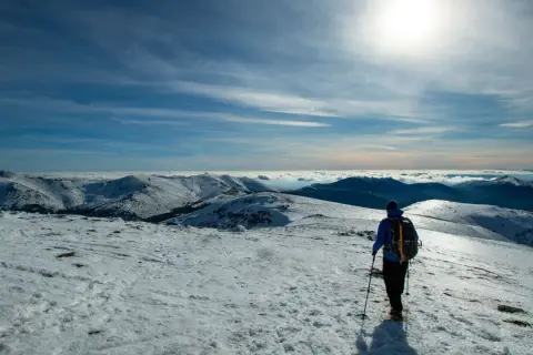 Senderismo en la nieve de Peñalara. Un excursionista con mochila y bastones contempla un paisaje montañoso cubierto de nieve bajo un cielo azul.