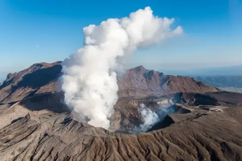 Fumée s'échappant du cratère du volcan Aso, sous un ciel bleu clair avec la lune visible.