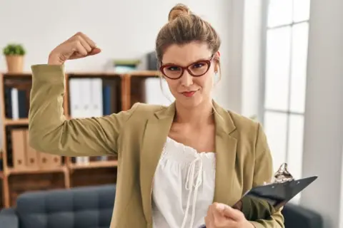Kobieta w okularach pokazuje biceps, symbolizując, jak być silnym psychicznie. W ręku trzyma teczkę.