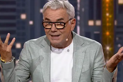 Mężczyzna w okularach i jasnej marynarce gestykuluje rękami na tle miejskiego krajobrazu.