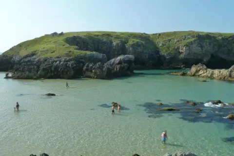 Gente disfrutando del agua cristalina en la costa de Llanes, con acantilados verdes y rocosos de fondo.