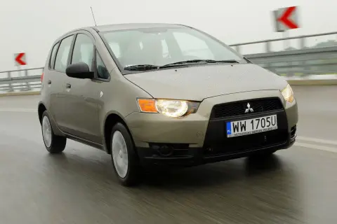 Złoty Mitsubishi Colt 2006 na mokrej drodze, światła włączone, w tle znaki drogowe.