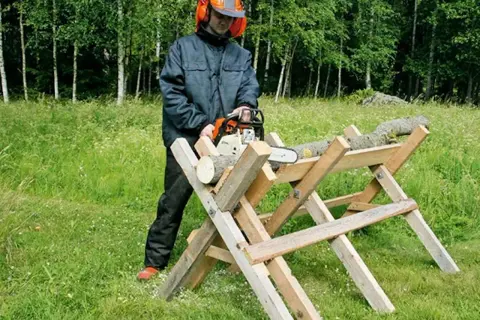 Man med motorsåg använder en hemmagjord sågbock för att kapa ved i en grönskande skogsmiljö.