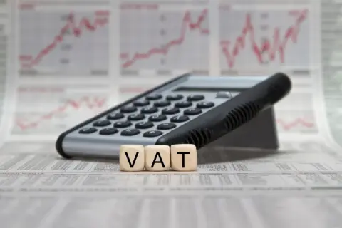 Kalkulator i kostki z literami VAT na tle wykresów finansowych. Pełna księgowość krok po kroku.