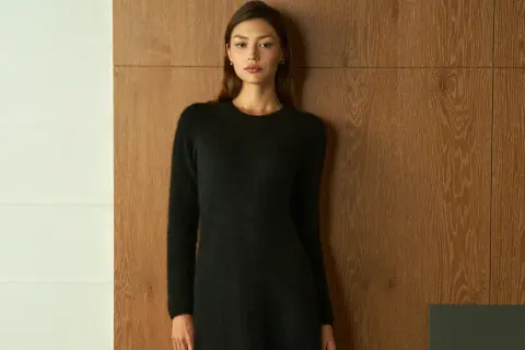Frau in schwarzem Strickkleid vor Holzvertäfelung. Ein schlichtes Outfit, das zu den modernen Dresscodes für Frauen passt.