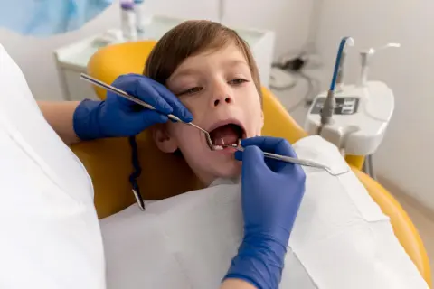 Chłopiec przechodzi leczenie amputacyjne zębów mlecznych. Dentysta w niebieskich rękawiczkach bada jego ząbki.