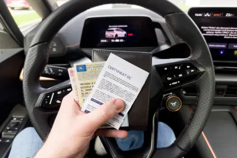 Kierowca trzyma dowód rejestracyjny i certyfikat ubezpieczenia OC na dzień w dłoni, siedząc za kierownicą nowoczesnego samochodu.