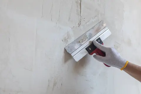 Ręka w rękawicy wykonuje szpachlowanie ścian, wygładzając nierówności.
