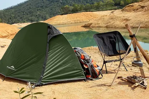 Tienda de campaña verde junto a un lago, con una silla plegable negra y mochilas de transporte.