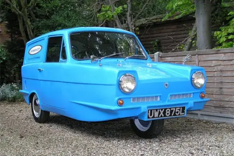En blå Reliant bil, en trehjulig klassiker, står parkerad på grus.