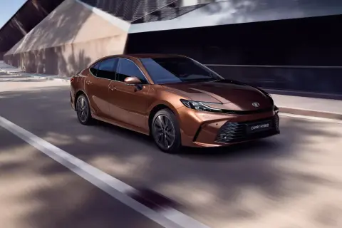 Brązowa Toyota Camry Hybrid 2024 pędzi po drodze, otoczona nowoczesną architekturą i cieniami drzew.
