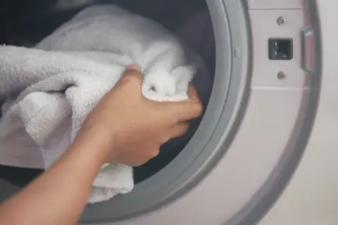 Ręka wkłada białe ręczniki do pralki. Dowiedz się, jak wyprać mocno zabrudzone ręczniki, aby znów były jak nowe.
