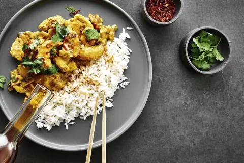 Ein Teller mit Reis und einem köstlichen Thai Curry, garniert mit frischem Koriander. Daneben kleine Schälchen mit Chiliflocken und Koriander.