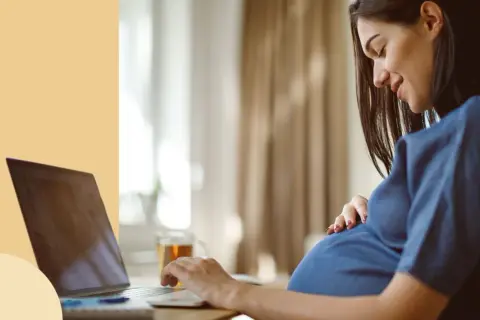 Ciężarna kobieta pracuje przy laptopie, czując ruchy dziecka. Ułożenie główkowe i aktywność malucha są widoczne.