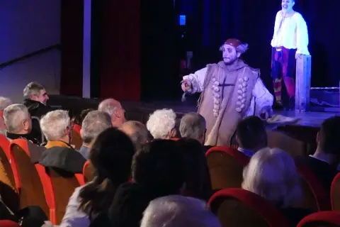 Attori sul palco, uno con un costume particolare, interagiscono con il pubblico, rompendo la quarta parete.