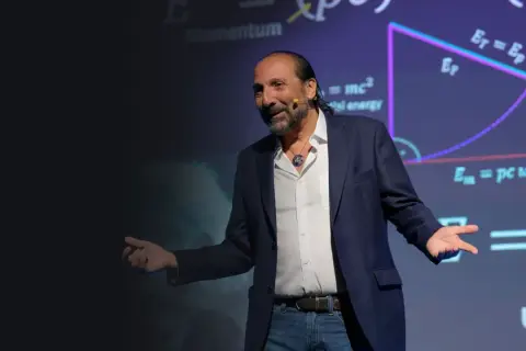 Nassim Haramein, con gestos amplios, explica conceptos de física ante una pantalla con fórmulas como E=mc².