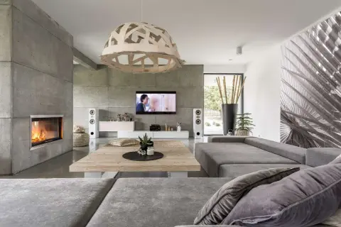 Nowoczesny salon z kominkiem i ścianą z betonu architektonicznego. Szara kanapa, drewniany stolik i designerska lampa tworzą przytulną atmosferę.