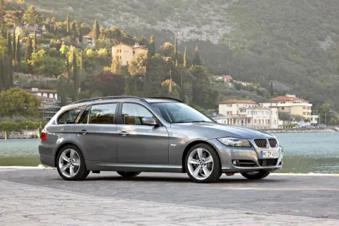 Szary kombi BMW 325i zaparkowany nad jeziorem, z malowniczymi wzgórzami i budynkami w tle.