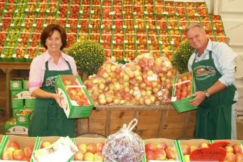 Ein Paar präsentiert Äpfel auf dem markt leichlingen. Sie halten Kisten mit frischen Äpfeln, umgeben von weiteren Obstkisten.