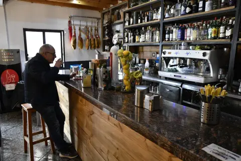 Hombre en taburete en bar Almería Enix, con jamones colgados y estanterías llenas de licores.