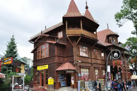 Drewniana architektura na Krupówkach, z charakterystycznymi zdobieniami i wieżyczkami. W tle widać ludzi spacerujących po tętniącej życiem ulicy.