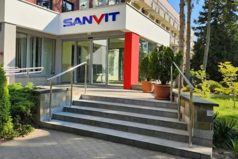 Wejście do sanatorium Sanvit w Ciechocinku. Schody prowadzą do przeszklonych drzwi, a nad nimi widnieje niebiesko-czerwone logo.