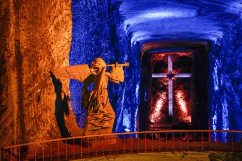 Statua di un angelo con tromba nella cattedrale del sale, illuminata da luci blu e arancioni, con una croce sullo sfondo.
