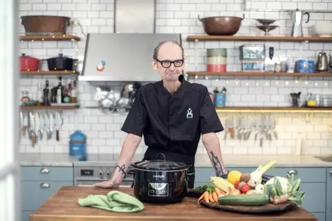 En kock med tatueringar står vid ett köksbord med en slow cooker och färska grönsaker. Perfekt för att hitta nya ångkokare recept.