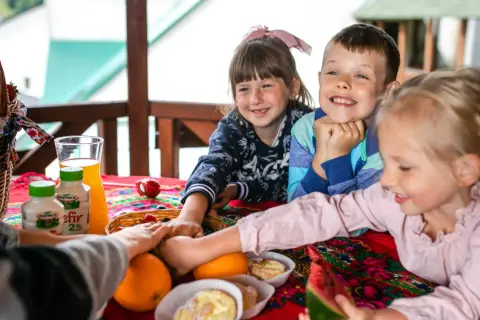 Radosna majówka w górach z dziećmi. Uśmiechnięte maluchy przy stole pełnym owoców, kefirów i słodkości.