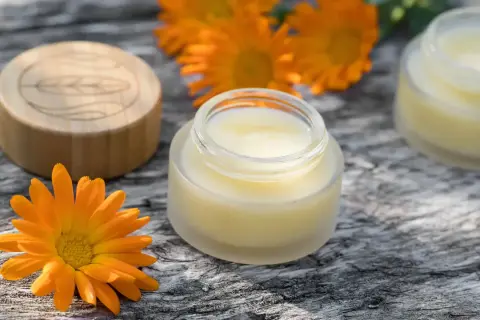Ringelblume-Salbe in Gläschen, umgeben von Blüten. Die Ringelblume wirkt beruhigend auf die Haut.