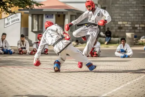 Två unga män i vit kampsportsdräkt, med skydd, utövar kampsport med pinnar.