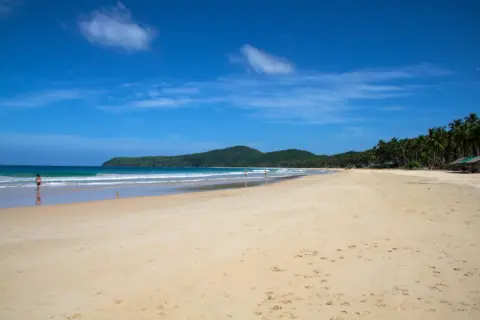 Słoneczny Nacpan Beach z turkusową wodą, zielonymi wzgórzami i palmami. Kilka osób spaceruje po piaszczystym brzegu.