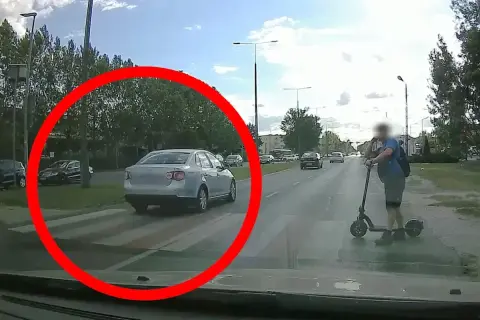 Srebrny samochód na pasach, wymuszenie pierwszeństwa. Na hulajnodze stoi mężczyzna.